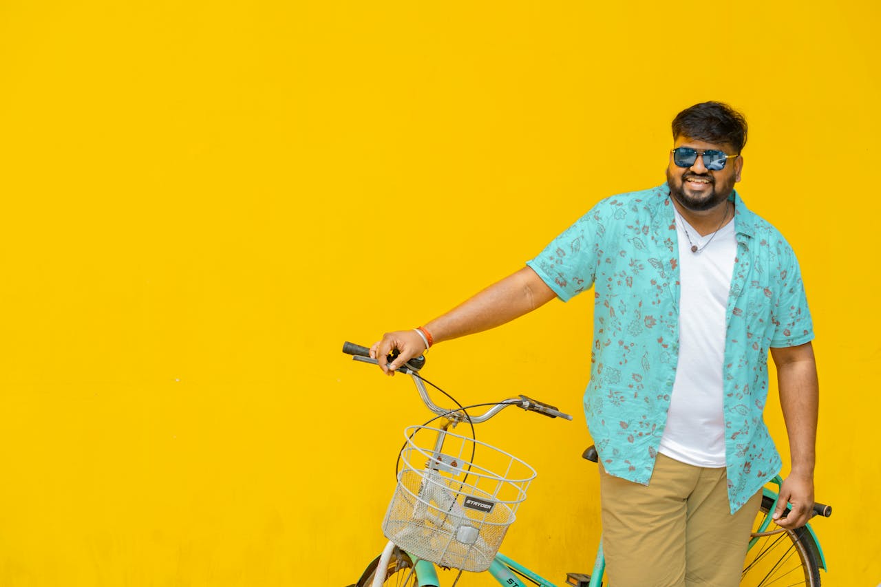 About Cheerful man in colorful attire posing with a bicycle against a bright yellow wall.